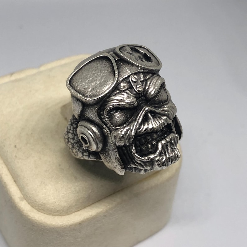 K&ouml;nnte beinhalten: Ein silberfarbener Ring mit einem detaillierten Totenkopf-Design, der eine Fliegerbrille tr&auml;gt. Der Sch&auml;del hat einen bedrohlichen Ausdruck mit sichtbaren Z&auml;hnen. Der Ring ist wahrscheinlich aus Metall und ein einzigartiges Schmuckst&uuml;ck.