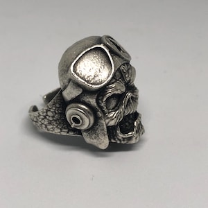 K&ouml;nnte beinhalten: Ein silberfarbener Ring mit einem Totenkopf, der einen Pilotenhelm und eine Brille tr&auml;gt. Der Ring weist aufwendige Details auf, darunter die Gesichtsz&uuml;ge des Sch&auml;dels und die Textur des Helms. Der Ring ist zum Tragen am Finger konzipiert.
