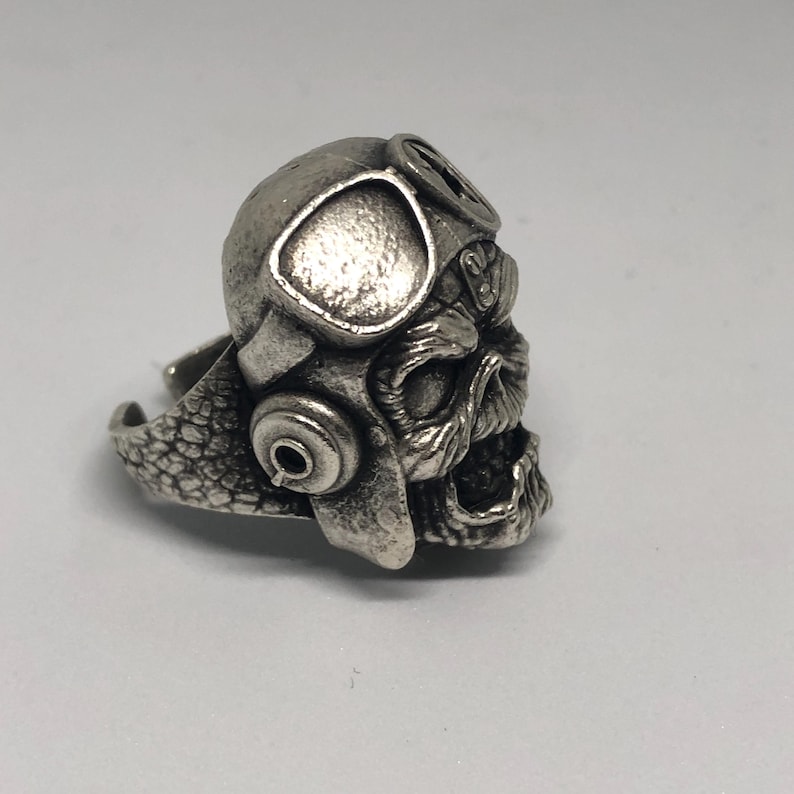 K&ouml;nnte beinhalten: Ein silberfarbener Ring mit einem Totenkopf, der einen Pilotenhelm und eine Brille tr&auml;gt. Der Ring weist aufwendige Details auf, darunter die Gesichtsz&uuml;ge des Sch&auml;dels und die Textur des Helms. Der Ring ist zum Tragen am Finger konzipiert.