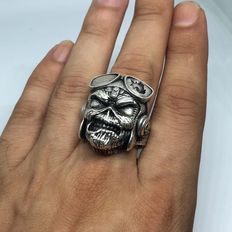K&ouml;nnte beinhalten: Ein silberner Ring mit einem Totenkopf-Design, komplett mit Fliegerbrille und Kopfh&ouml;rern. Der Totenkopf hat detaillierte Merkmale und ist das zentrale Element. Der Ring wird an einem Finger getragen.