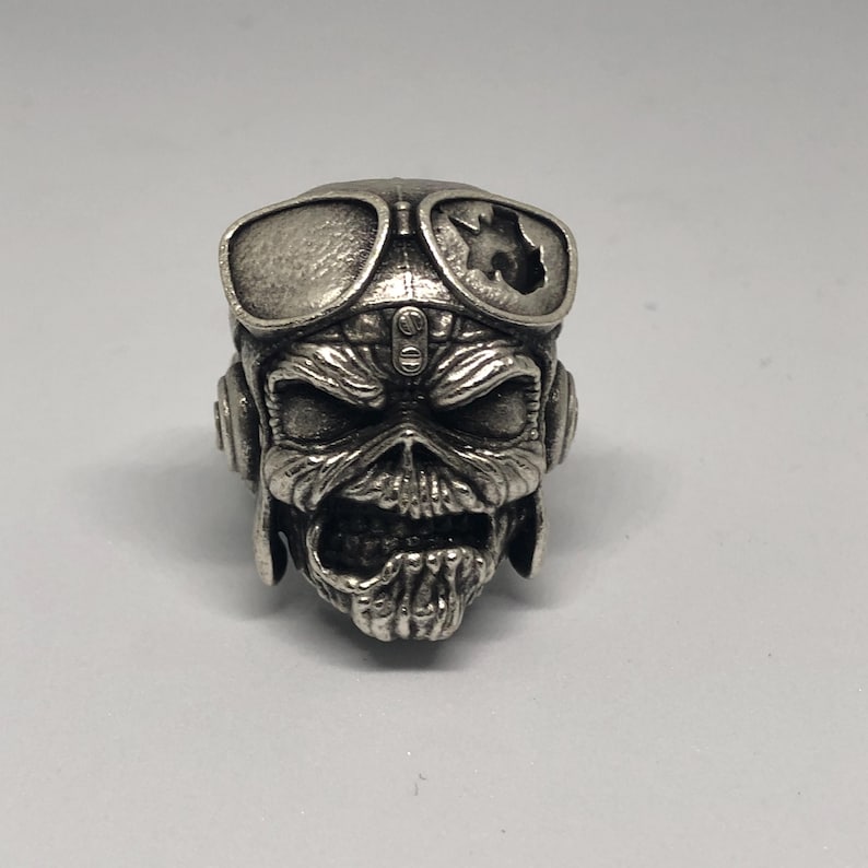 K&ouml;nnte beinhalten: Ein silberner Ring mit einem detaillierten Totenkopf-Design, der eine Fliegerbrille tr&auml;gt. Der Sch&auml;del hat einen bedrohlichen Ausdruck mit sichtbaren Z&auml;hnen und Augenh&ouml;hlen. Die Brille hat eine zerbrochene Linse, was dem Ring eine einzigartige, kantige &Auml;sthetik verleiht.
