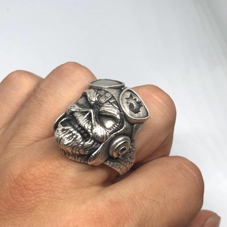 K&ouml;nnte beinhalten: Ein silberfarbener Ring mit einem detaillierten Sch&auml;deldesign, Helm und Seitenemblem. Der Ring wird am Finger getragen und zeigt die aufwendige Handwerkskunst und das gotische Thema.