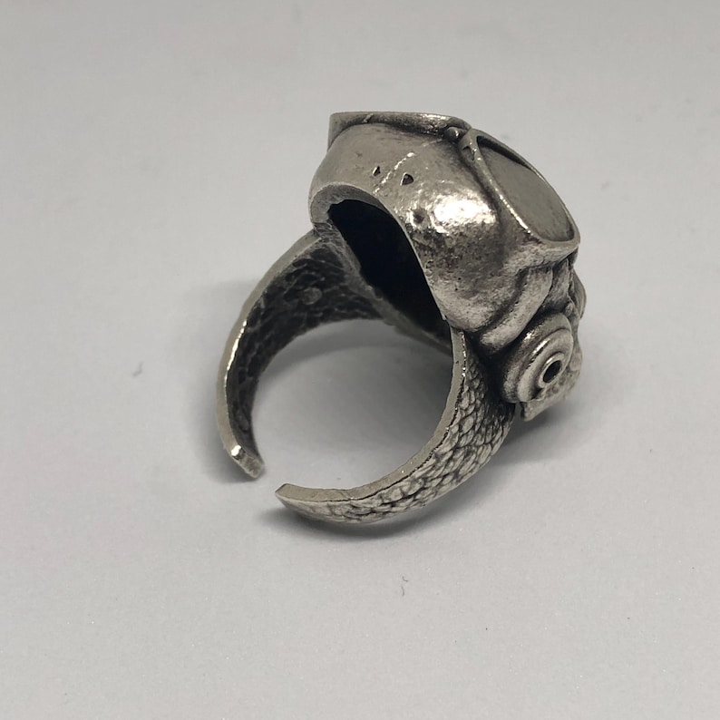K&ouml;nnte beinhalten: Ein silberfarbener Ring mit einem einzigartigen Design. Der Ring hat eine detaillierte, strukturierte Oberfl&auml;che mit offenem Design. Das Design des Rings umfasst eine abgerundete Oberseite mit einem kreisf&ouml;rmigen Detail und eine schuppenartige Textur am Band.