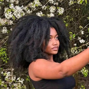 May include: A person with dark skin and voluminous black hair poses outdoors. She wears a black tank top, with a backdrop of white flowers and green foliage. The image is well-lit, highlighting her features and the natural setting.