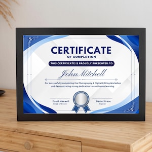 May include: A framed certificate of completion in a black frame. The certificate features a blue and white design with the text "CERTIFICATE OF COMPLETION" and "John Mitchell". It rests on a wooden surface.