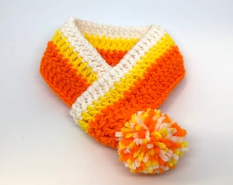 May include: A crocheted scarf in candy corn colours: orange, yellow, and white stripes. It has a matching pom-pom. The scarf is designed to be worn around the neck, a cozy accessory.
