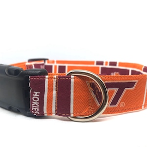 May include: Dog collar featuring the Virginia Tech Hokies logo in orange and maroon. The collar has a black plastic buckle and a silver metal D-ring. The fabric displays the team's logo and the word "Hokies".