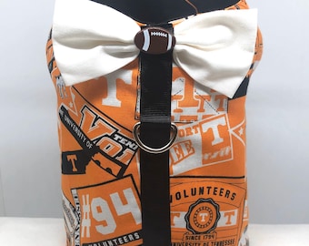 Könnte beinhalten: Orange-weißes Hundegeschirr mit weißer Schleife und footballförmigem Knopf. Das Geschirr zeigt ein sich wiederholendes Muster aus Logos und Text der Tennessee Volunteers. Ein schwarzer Riemen mit einem D-Ring aus Metall ist angebracht.