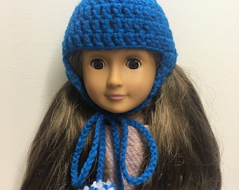 May include: A blue crochet hat with a white and blue pom-pom on top and two white and blue pom-poms at the ends of the ties. The hat is worn by a doll with long brown hair.