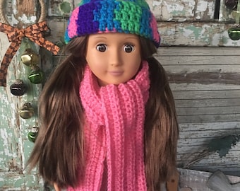 May include: A doll wearing a colourful crochet hat in shades of pink, blue, green, and purple, paired with a bright pink crochet scarf. The hat and scarf are handmade, showcasing a cosy, textured design. The doll has long brown hair.