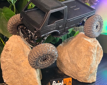 May include: A black 3D printed pickup truck model with a flatbed, sitting on two rocks. The truck has a detailed chassis and wheels. The truck is printed by PrintHub.