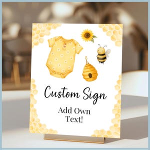 May include: A custom sign with a yellow and white watercolor design. The sign features a yellow baby onesie, a sunflower, a bee, and a beehive. The text on the sign reads "Custom Sign" and "Add Own Text!"