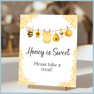Puede incluir: Un letrero blanco con soporte dorado presenta un diseño de acuarela con una abeja, girasol, body de bebé, colmena y sol. El texto dice: "Honey is Sweet, Please take a treat!" Los bordes de panal enmarcan el letrero.