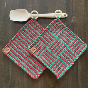 May include: Two red and green striped pot holders with leather tags that say "Happy Home" and "To Knit". The pot holders are hanging on a white spatula with two wooden rings.