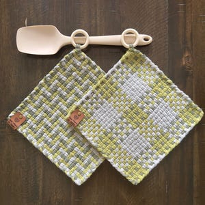 May include: A woven kitchen set featuring two square potholders in a green, gray, and white pattern, along with a matching beige spoon and a hanging bar. The potholders have leather tags. The set is designed for kitchen use.