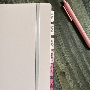 May include: A pale pink planner with a matching pen. The planner features a grey elastic band and pink tabs. Tabs are labelled with terms such as "To Do", "Budget", "Renos", "Repairs", "Wishlist", and "Goals".
