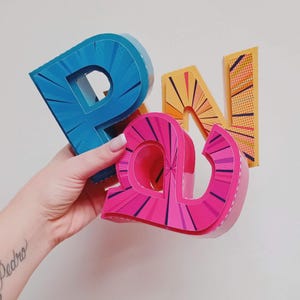Puede incluir: Tres letras de cartón de colores que deletrean "PAN" en estilo cómic. Las letras son azules, rosas y amarillas con fondo blanco y contornos negros.