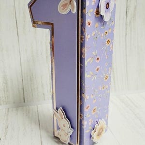 Puede incluir: Una caja de color morado en forma de número uno con un patrón floral. La caja tiene un adorno de mariposa, un conejito y una flor.