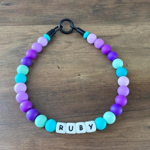 Pode incluir: Um colar de contas com um fecho preto e o nome "RUBY" escrito com contas de letras brancas. O colar apresenta um gradiente de contas coloridas, incluindo roxo, turquesa e lilás, criando um acessório vibrante.