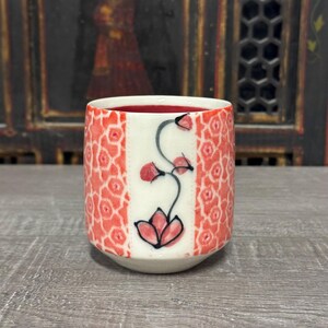 May include: A ceramic cup with a red floral pattern and a white vertical stripe featuring a stylized flower design. The cup's interior is red, and the exterior has a repeating geometric pattern in red.