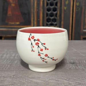May include: A white ceramic bowl with a red interior and a floral design. The exterior features a delicate black and red branch pattern. The bowl has a rounded shape and a small, raised base, suitable for serving or display.