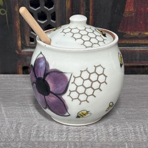 Può includere: Un vasetto per miele in ceramica bianca con coperchio abbinato e un mestolo in legno. Il vasetto presenta un fiore viola dipinto a mano, motivi a nido d'ape marroni e piccole api gialle e nere sia sul corpo che sul coperchio.