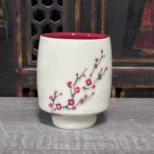 Puede incluir: Una taza cilíndrica de cerámica con exterior de color crema que presenta un diseño pintado a mano de una rama de flor de ciruelo en rojo y negro. El interior de la taza es de un color rojo intenso sólido. Acabado liso y brillante.