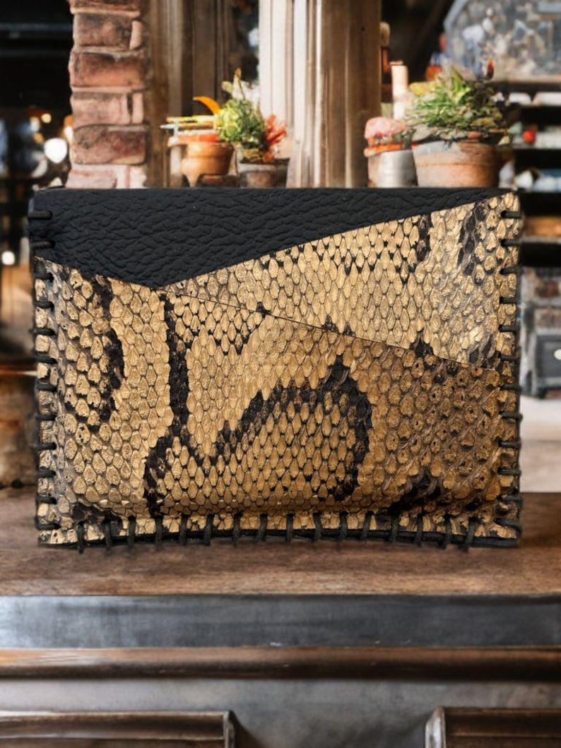 Pu&ograve; includere: Una piccola pochette in pelle di serpente nera e dorata con un bordo cucito.