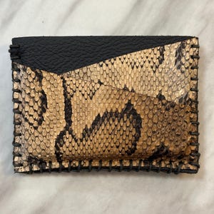 Pu&ograve; includere: Un piccolo portafoglio in pelle di serpente nero e oro con un bordo cucito.