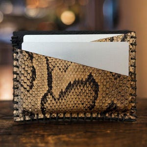 Pu&ograve; includere: Un piccolo portafoglio in pelle nera con un motivo a pelle di serpente e un porta carte argentato. Il portafoglio ha un bordo cucito.