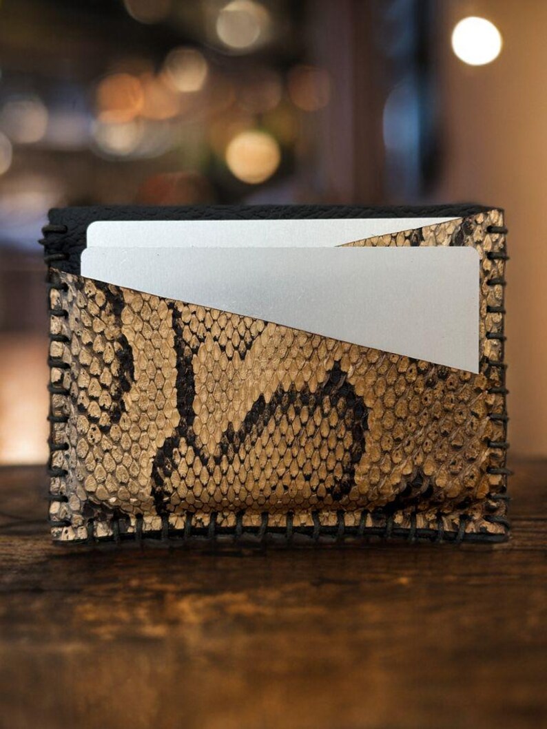 Pu&ograve; includere: Un piccolo portafoglio in pelle nera con un motivo a pelle di serpente e un porta carte argentato. Il portafoglio ha un bordo cucito.