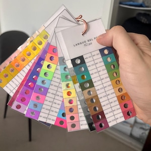 May include: A hand holds a fan of gel polish colour swatches. The swatches are arranged in a rainbow of colours, with each swatch labelled with a code. The text "LANGUO GEL POLISH 162 pcs" is printed on one of the cards.