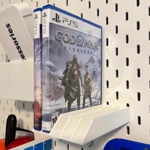 Puede incluir: Una caja de videojuego PS5 para God of War Ragnarok, con dos personajes con armas en un paisaje nevado. La caja del juego se muestra en un estante blanco, con otros accesorios visibles.