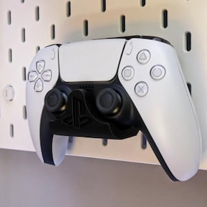 May include: A white and black PlayStation 5 controller mounted on a white pegboard. The controller has a symmetrical design with black joysticks and buttons, and a black holder. The pegboard has evenly spaced holes.