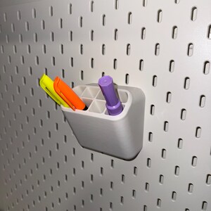May include: A white plastic organiser with six compartments holds three markers: one yellow, one orange, and one purple. The organiser is mounted on a white pegboard wall.