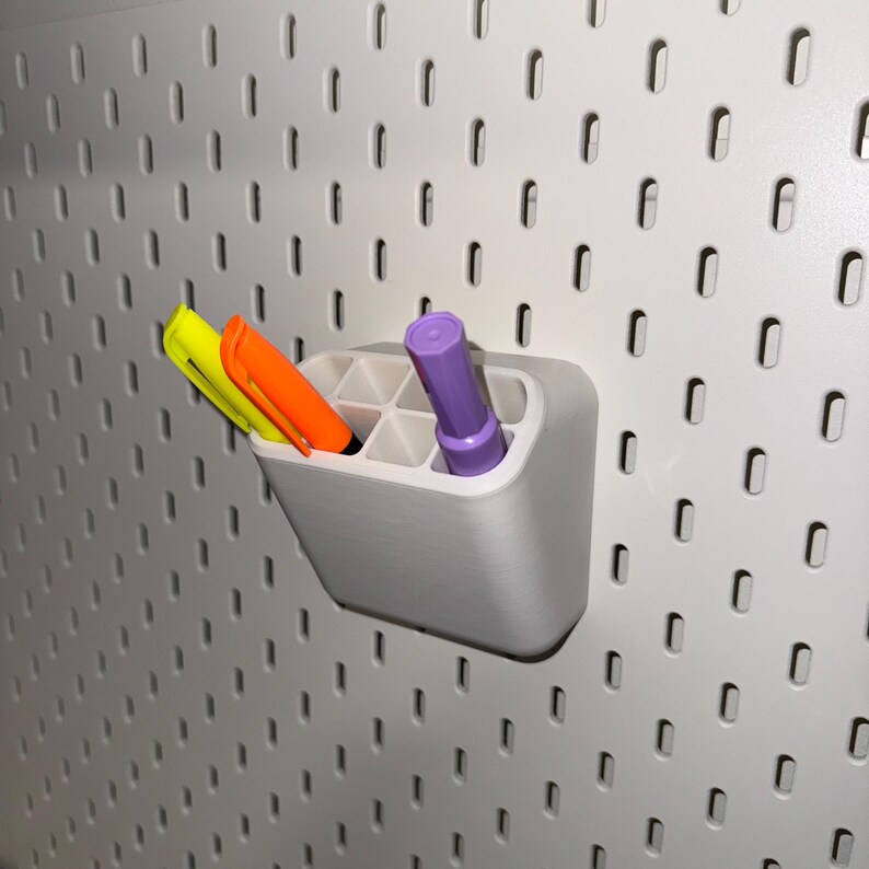 May include: A white plastic organiser with six compartments holds three markers: one yellow, one orange, and one purple. The organiser is mounted on a white pegboard wall.