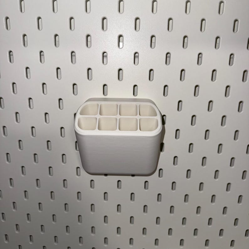 May include: White plastic organiser with 10 compartments, designed to hold small items like pens, pencils, or makeup brushes. The organiser is mounted on a pegboard wall.