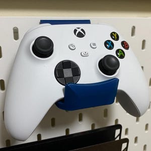 Peut inclure: Manette Xbox blanche fixée sur un panneau perforé blanc avec un support bleu. La manette a des joysticks noirs et des boutons colorés. Le panneau perforé a des trous espacés.