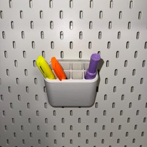 May include: A white plastic wall-mounted organiser with nine compartments, holding three markers: yellow, orange, and purple.