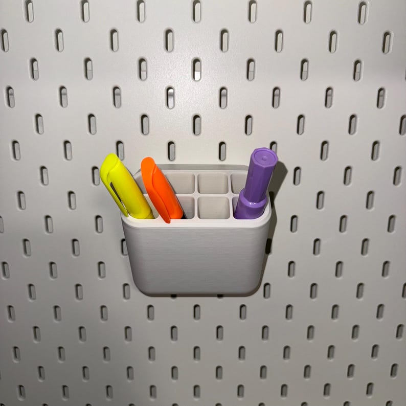 May include: A white plastic wall-mounted organiser with nine compartments, holding three markers: yellow, orange, and purple.