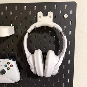 May include: White headphones hanging on a black pegboard wall with a white hook. The headphones are over-ear and have a white headband.
