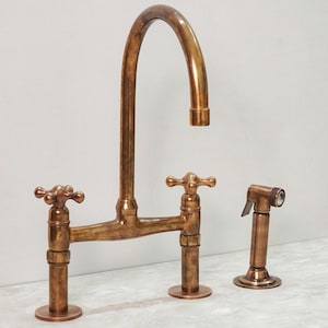 Op de afbeelding: Een bronzen keukenkraan set met een hoge booguitloop, twee kruisgreepventielen en een zijsproeier. De kraan heeft een vintage uitstraling met een warme, metallic afwerking. De set is ontworpen voor installatie op het aanrecht.