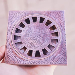 May include: A square, copper-colored drain cover with a decorative, circular grate design. The cover has a hammered texture.