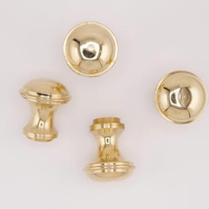 Könnte beinhalten: Vier polierte Messing-Schrankknöpfe und -Griffe. Zwei sind abgerundet, zwei sind zylindrisch mit dekorativen Details. Die Hardware hat eine leuchtend goldene Farbe und reflektiert das Licht. Die Knöpfe sind wahrscheinlich für Schubladen oder Schränke.
