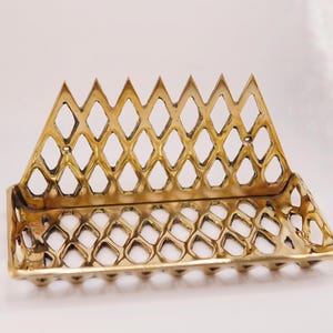 Puede incluir: Una bandeja decorativa de metal dorado con un diseño calado en forma de diamante. La bandeja tiene una base rectangular y una parte trasera triangular, creando una pieza de exhibición única. La bandeja probablemente se utiliza para guardar objetos pequeños.