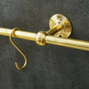 Könnte beinhalten: Ein Küchenutensilienhalter aus Messing mit einer runden, dekorativen Endkappe und vier Haken. Der Halter ist an einer Wand montiert.
