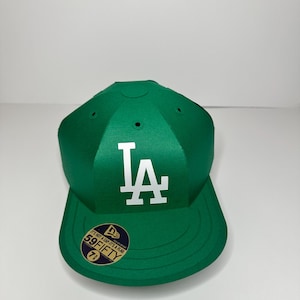 May include: A green baseball cap with a white "LA" logo on the front. The cap has a black New Era logo and a sticker that says "It's not a cap, it's a flag 59FIFTY 7 1/4".