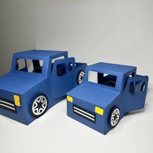 May include: Two blue paper model pickup trucks with open doors. The trucks have black and white wheels, black and white front bumpers, and yellow headlights. The trucks are on a white surface.