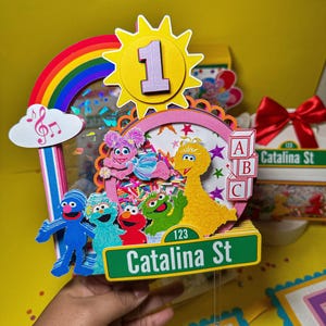 K&ouml;nnte beinhalten: Ein farbenfroher Tortenaufsatz zum Geburtstag mit Sesamstra&szlig;en-Charakteren, einem Regenbogen und der Zahl 1. Der Aufsatz enth&auml;lt ein Stra&szlig;enschild mit der Aufschrift "Catalina St" und ABC-Bl&ouml;cke. Das Design ist lebendig mit verschiedenen Farben.