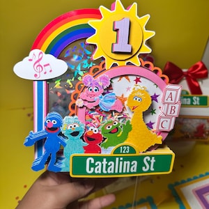 K&ouml;nnte beinhalten: Ein farbenfroher Tortenaufsatz zum Geburtstag mit Sesamstra&szlig;en-Figuren, einem Regenbogen und einer Sonne mit der Zahl 1. Der Aufsatz enth&auml;lt ein Stra&szlig;enschild mit der Aufschrift "Catalina St". Das Design ist lebendig mit Rosa, Gelb, Blau und Gr&uuml;n.